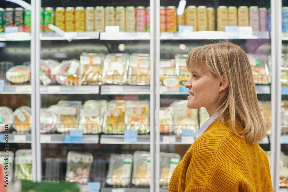 Side view portrait of blonde adult woman walking by freeze shelves in supermarket while grocery shopping, copy space