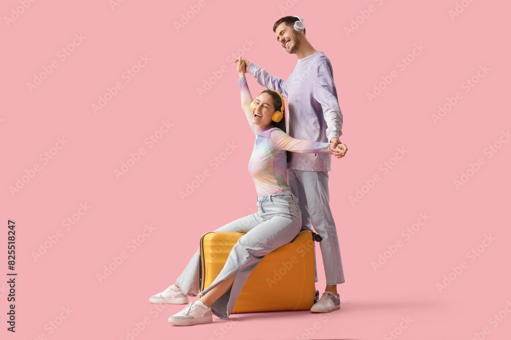 Fototapeta premium Beautiful young happy couple of tourists in headphones with suitcase listening music on pink background