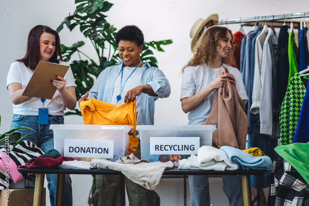 Obraz premium Concept of textile pollution, conscious consumption. Ecology, sustainable lifestyle. Group of volunteers, young women, sorting clothes in charitable foundation for charity donation, recycling.