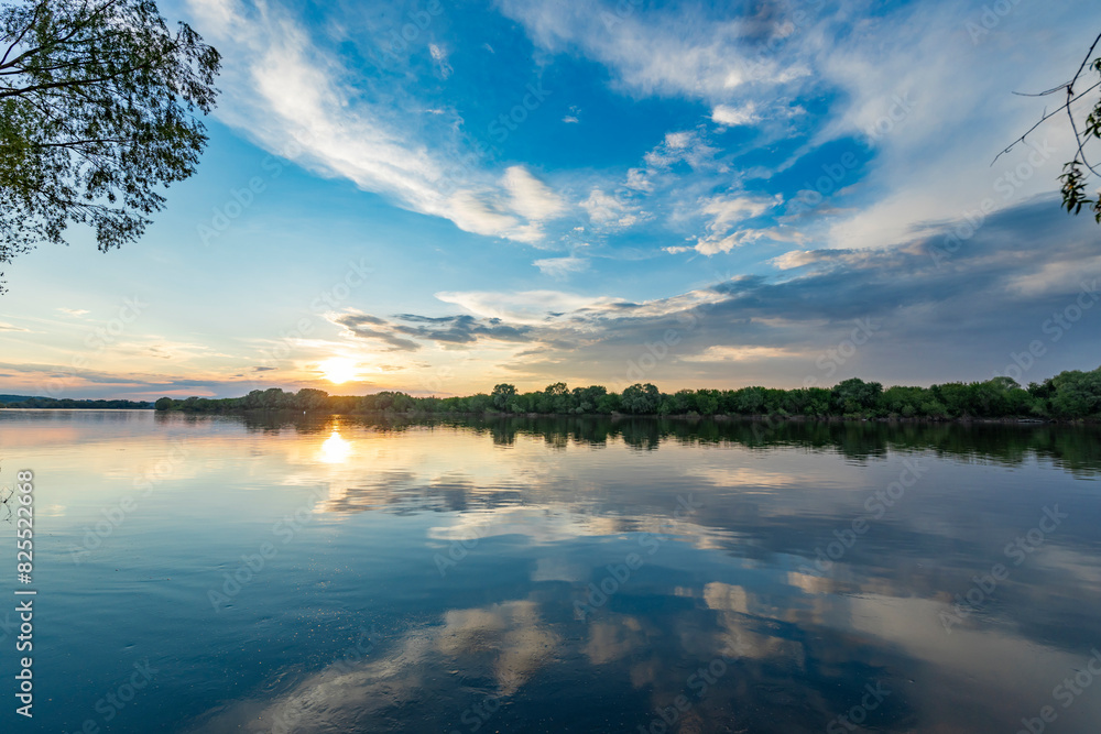 Obraz premium Vibrant hues of orange and gold paint the sky as the sun sets over the tranquil waters of a springtime lake.