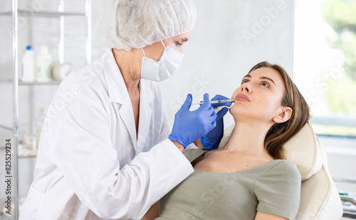 Hands of cosmetologist making filler injection in female lips. Young woman is receiving procedure with enjoy.