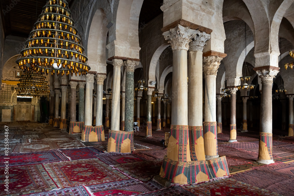 Obraz premium Empty grand prayer hall of Great Mosque of Kairouan with rows of Corinthian columns, hanging chandeliers, and traditional carpets, Tunisia