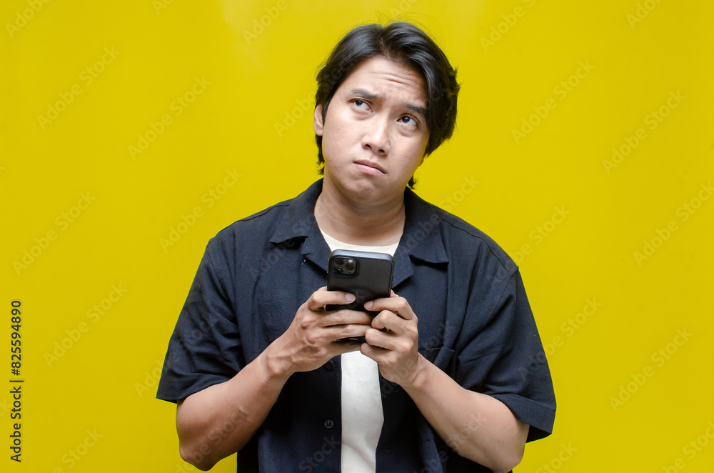 an overthinker young Asian man in pensive mood holding phone with sad and thoughtful expression. Asian man holding phone while questioning and wondering. an overthinker young Asian man in pensive mood holding phone with sad and thoughtful expression. Asian man holding phone while questioning and wondering.