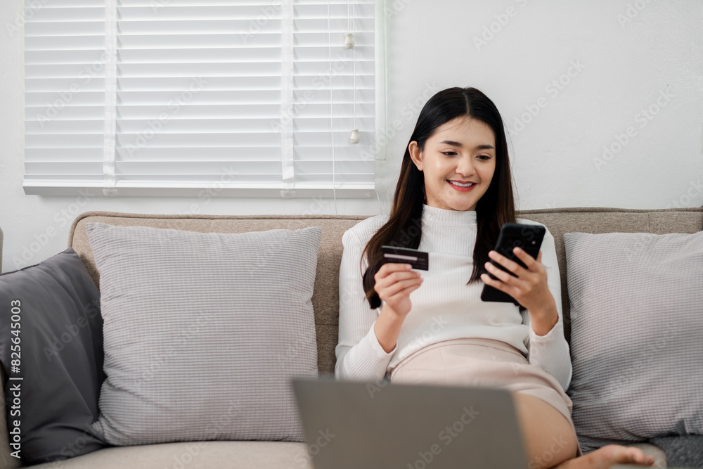 A woman is sitting on a couch and looking at her phone. She is holding a credit card and a laptop. Concept of relaxation and leisure, as the woman is using her phone and laptop to make a purchase