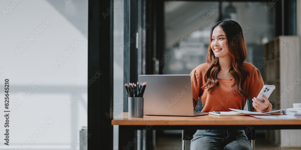 A woman is sitting at a desk with a laptop and a cell phone. She is smiling and she is happy A woman is sitting at a desk with a laptop and a cell phone. She is smiling and she is happy