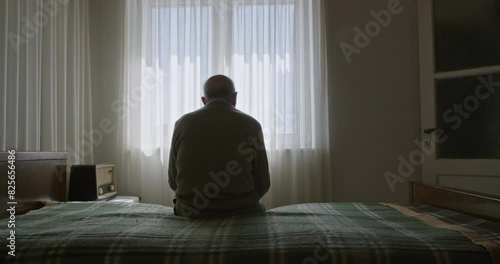 An elderly man sits alone on a bed in a sunlit room, staring out a window with sheer curtains.