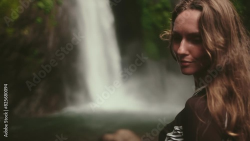 Woman standing in front of powerful Bali waterfall, surrounded by lush greenery. The water cascades down creating a dramatic backdrop for the serene moment. Air Terjun Munduk, Melanting. Slow motion photo