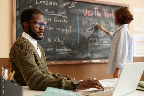 Selective focus shot of young African American teacher siting at desk using laptop while his student writing on blackboard