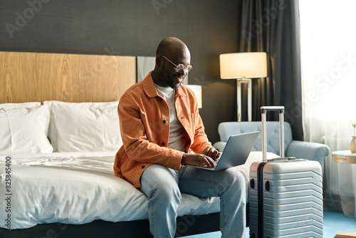 Fotografie Portrait of smiling African American man using laptop sitting on bed in hotel ro
