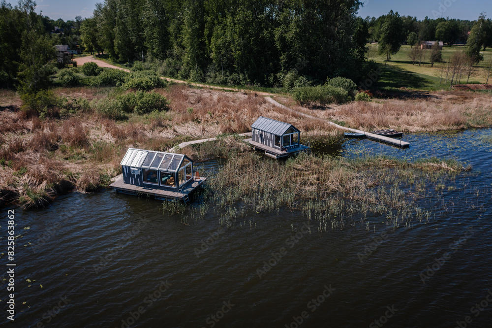 Two quaint floating houses rest on a serene lake surrounded by lush vegetation under a clear blue sky. Two quaint floating houses rest on a serene lake surrounded by lush vegetation under a clear blue sky.