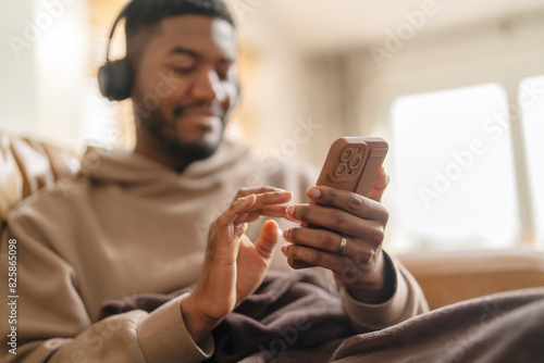 Happy man in headphones using phone, studying, learning, shopping, working online, listening to music, audiobook, podcast sitting on sofa at home 