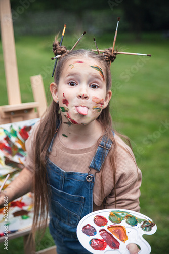 Portrait of a funny girl showing her tongue in paint on the background of green grass artist paints a picture