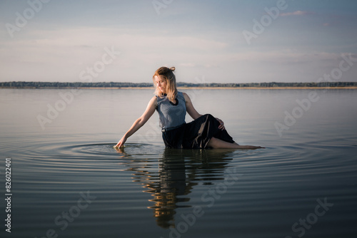 Portrait of beautiful young woman in dress sits in the water doing circles on water. Copy space. Fashion concept. Energy of water, earth and sun. Witch ritual. State of pleasure and bliss. Sexy pose