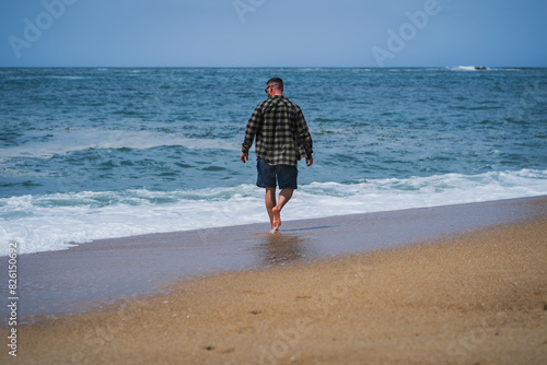 A man in a shirt and shorts walks barefoot along the beach in Nazaré on a summer day.