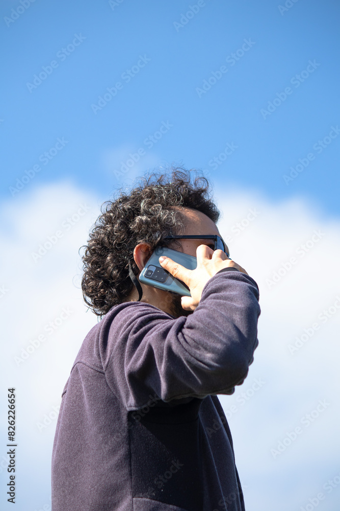 Fototapeta premium Turkish Man talking on mobile phone on blue sky background. Curly haired man with smartphone. 