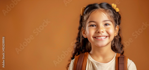 A young girl with brown hair and a brown backpack is smiling. She is wearing a white shirt and has her hair in pigtails. a joyful young  school aged hispanic girl wearing a backpack