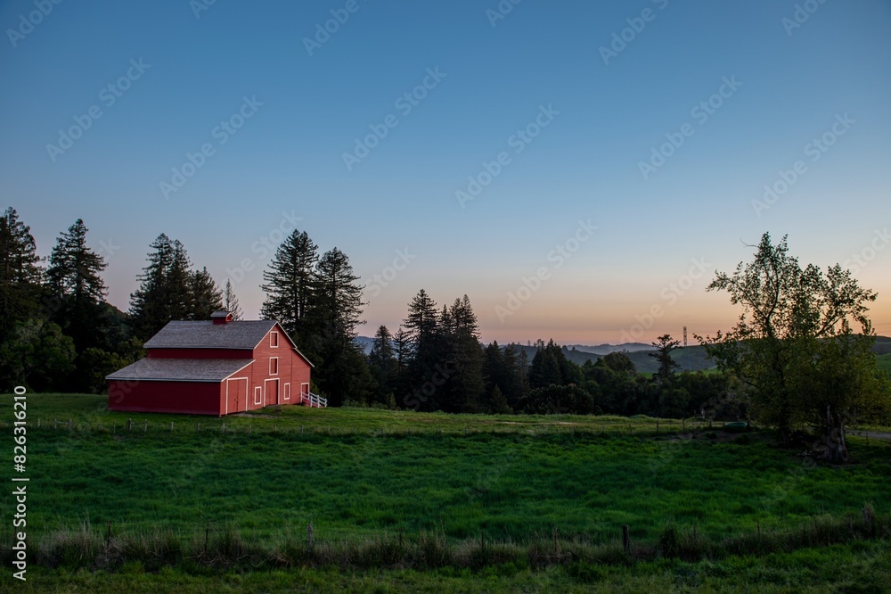 Red barn in the middle of a green field with trees, sky and landscape in the distance Red barn in the middle of a green field with trees, sky and landscape in the distance