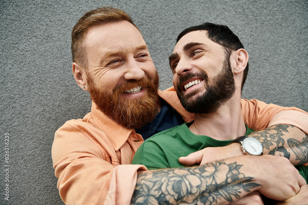 Two men with tattoos on arms, in casual attire, embrace in love, standing against grey wall. Two men with tattoos on arms, in casual attire, embrace in love, standing against grey wall.