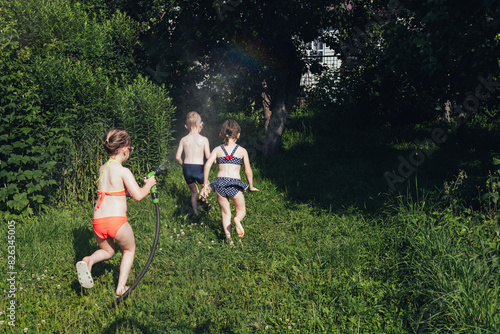 Two children run away from a girl with a hose. Children boy and girls in a swimsuit plays with splashes of water from a hose in the garden. Siblings, friends spend summer holidays, weekends together