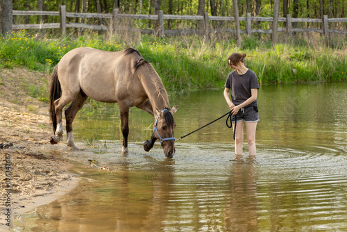A young woman practices bringing her horse into the water. The horse, trying to enter the pond, splashes water.