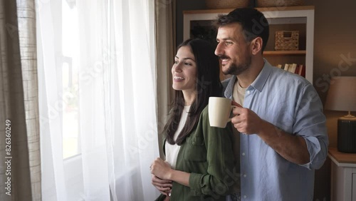 Young happy couple standing near window in their new home looking out, talking about neighbors, enjoying new life in quiet community. Man and woman enjoying new life at their new house.