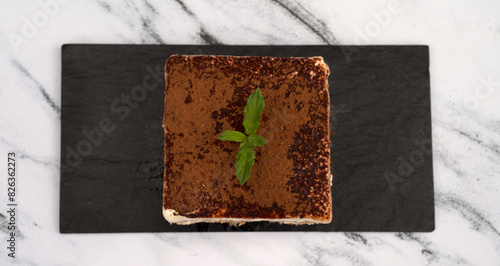 Sweet dessert gourmet presentation. Top view of a square slice of Tiramisu with chocolate covering on a black dish on the white marble table.