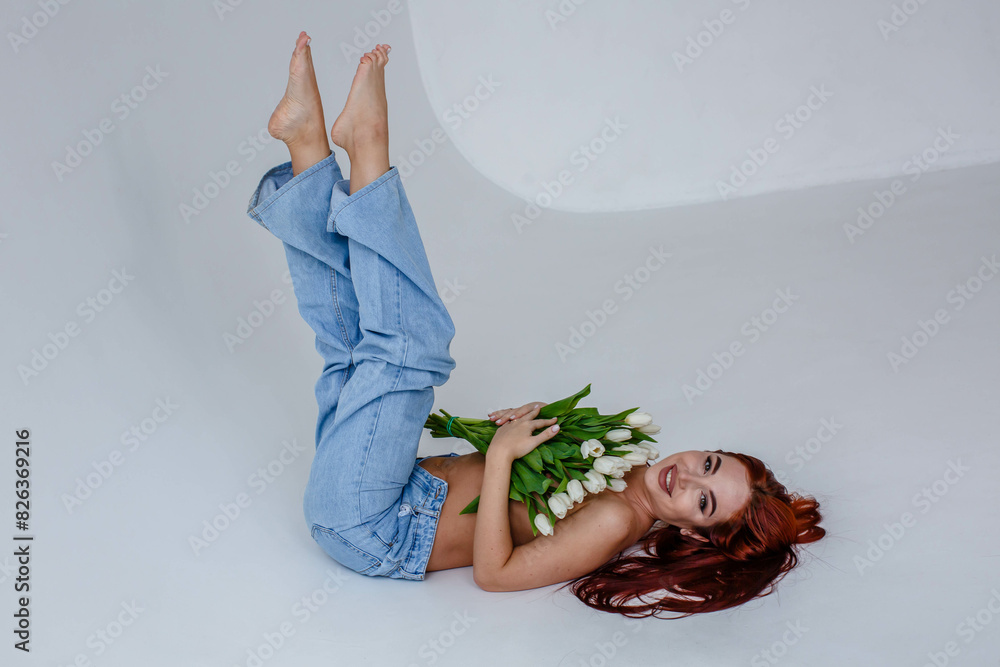 Red-haired girl in jeans with white tulips posing while lying on a white background