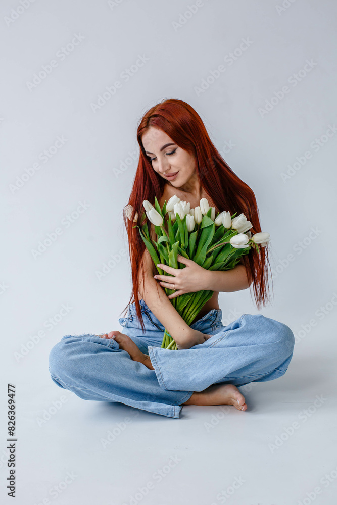 Red-haired girl in jeans with white tulips posing while sitting on a white background