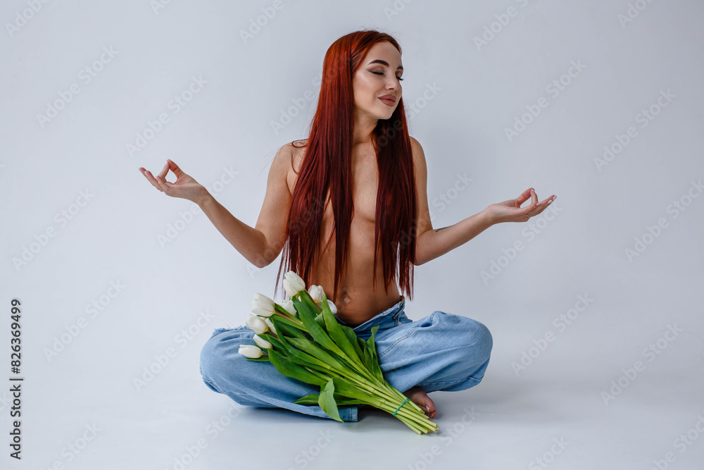 Red-haired girl in jeans with white tulips doing yoga while sitting on a white background