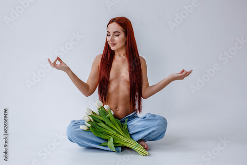 Red-haired girl in jeans with white tulips doing yoga while sitting on a white background