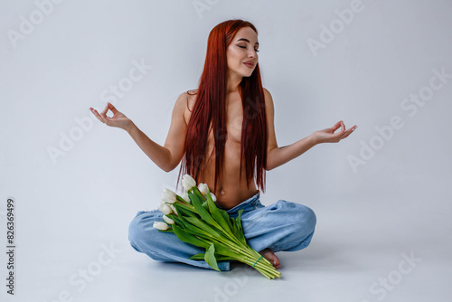 Red-haired girl in jeans with white tulips doing yoga while sitting on a white background