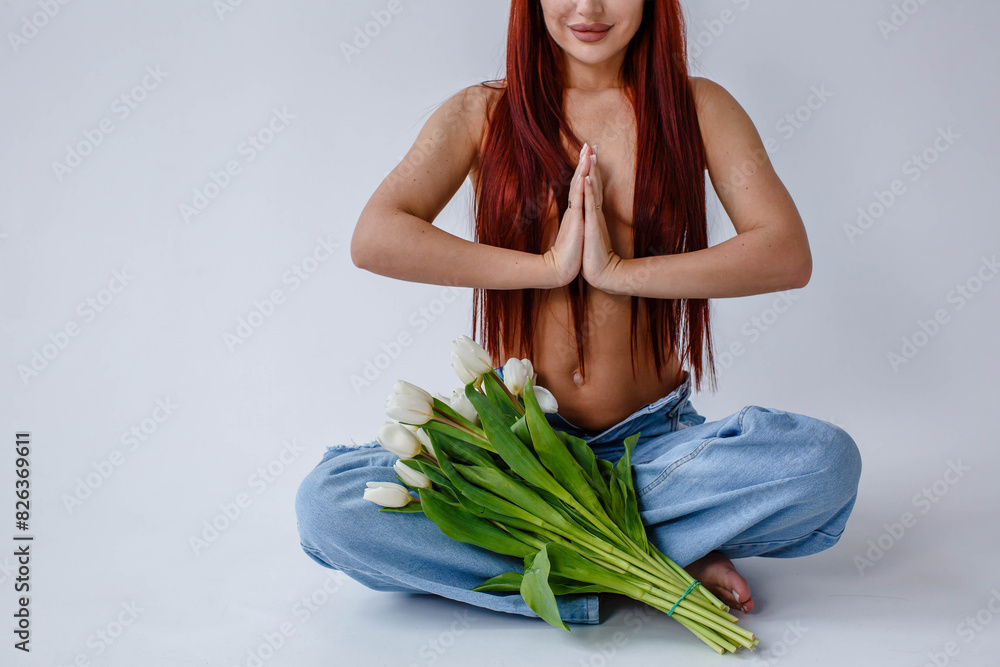 Red-haired girl in jeans with white tulips doing yoga while sitting on a white background
