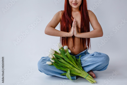 Red-haired girl in jeans with white tulips doing yoga while sitting on a white background