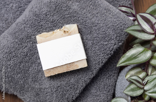 Soap bar with blank label on grey folded towel near green plant on wood in bathroom, mockup photo