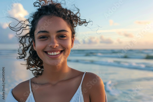 Portrait of young woman at sea looking at camera. Smiling latin hispanic girl standing at the beach with copy space and looking at camera. Happy mixed race girl in casual outfit with wind in her hair
