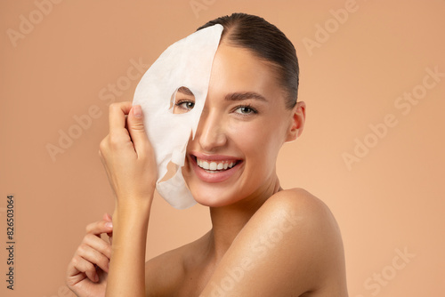 Young pretty European woman removing moisturizing, rejuvenating white fabric sheet mask from her face after using it, smiling at camera, beige background