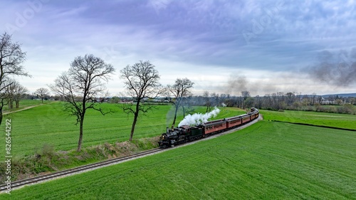 Принти на полотні Dramatic aerial capture of a vintage steam train chugging along a curvy track through lush green fields, with a backdrop of dramatic cloudy skies and a distant village