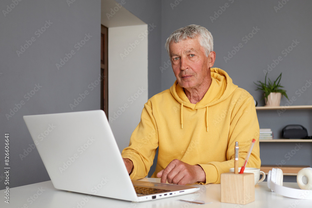 Gray haired mature man wearing casual yellow hoodie sitting at his home workplace typing on laptop keyboard looking at camera using computer for internet surfing Gray haired mature man wearing casual yellow hoodie sitting at his home workplace typing on laptop keyboard looking at camera using computer for internet surfing