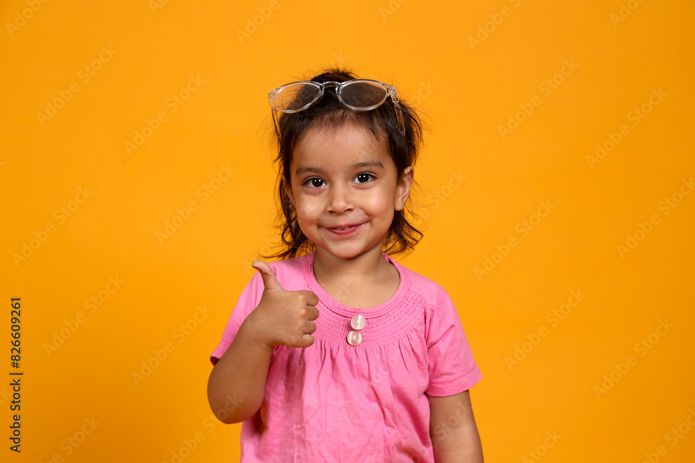 A young child with a confident smile. The child is wearing a pink shirt and large, clear-rimmed glasses. Showing thumbs up in a playful and self-assured manner A young child with a confident smile. The child is wearing a pink shirt and large, clear-rimmed glasses. Showing thumbs up in a playful and self-assured manner