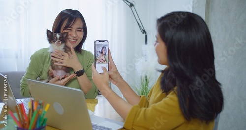 Two Asian women friend are sitting at a table with a small dog. women holds a Chihuahua and smiles for a selfie taking a picture of the dog with cell phone in a bright cozy workspace business home