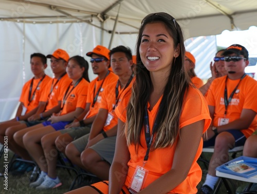 A group of people wearing orange shirts are sitting in a tent. A woman in an orange shirt is smiling at the camera photo