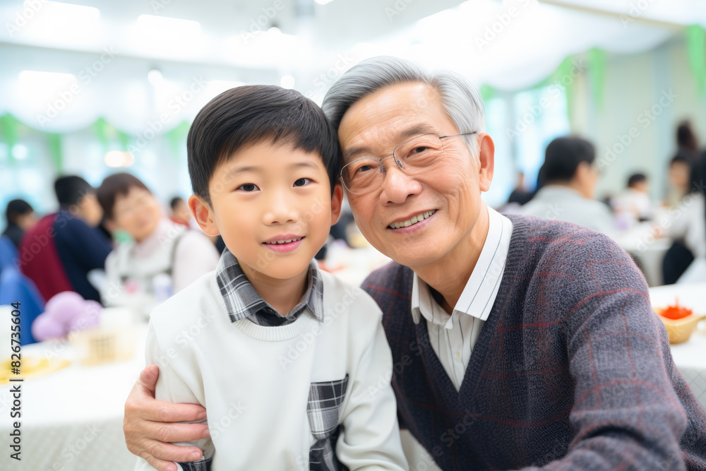 a touching moment captured between an older asian man and a young boy, likely his grandson, during a family celebration or gathering in a brightly lit, indoor setting. the pair are seated closely and a touching moment captured between an older asian man and a young boy, likely his grandson, during a family celebration or gathering in a brightly lit, indoor setting. the pair are seated closely and