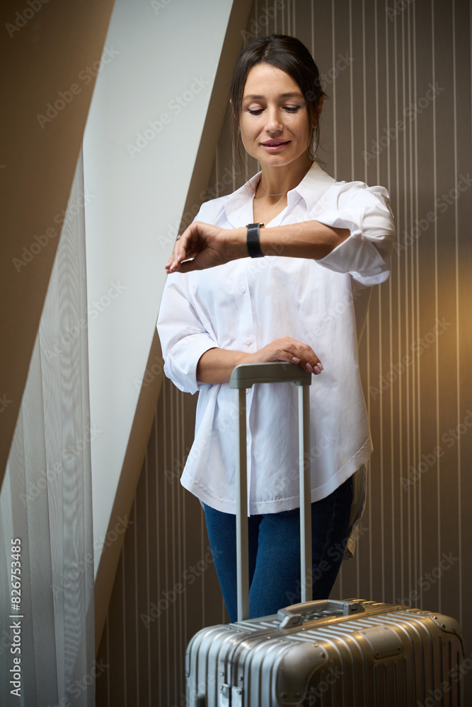 Front view of young woman with suitcase watching time on wristwatch by window Front view of young woman with suitcase watching time on wristwatch by window