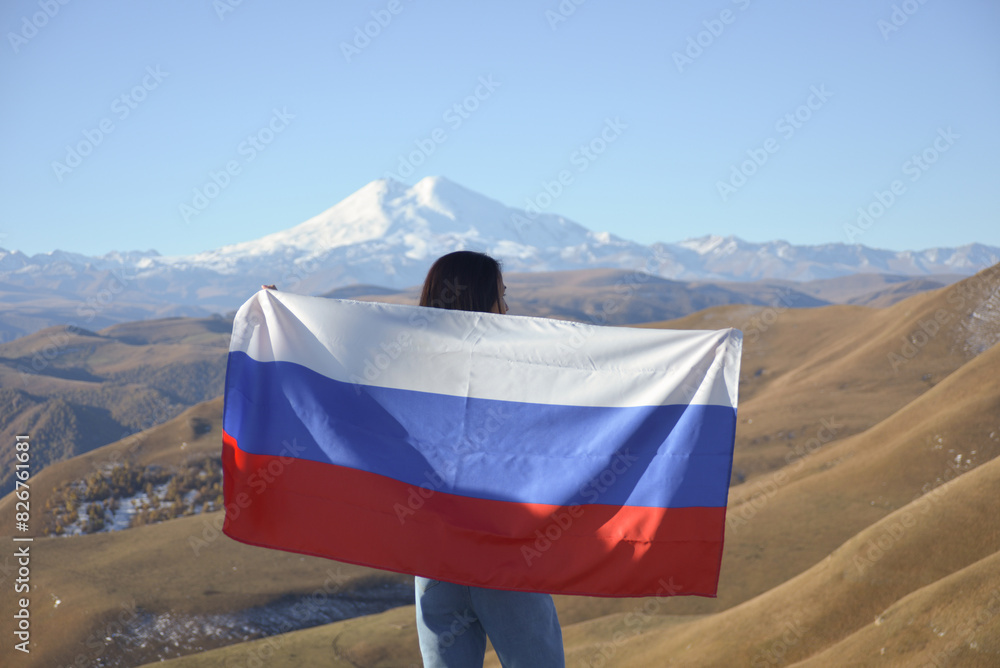 A young brunette woman stands against the backdrop of the snow-capped Mount Elbrus, looking at the beauty of the mountains, holding a Russian flag in her hands. Tricolor against the backdrop of snow