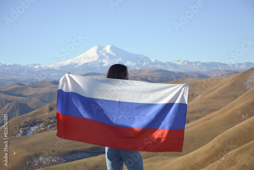 Obraz na płótnie A young brunette woman stands against the backdrop of the snow-capped Mount Elbrus, looking at the beauty of the mountains, holding a Russian flag in her hands. Tricolor against the backdrop of snow w salonie