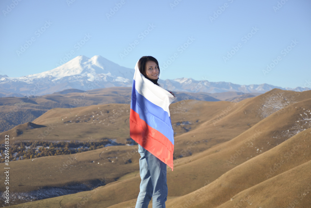 A young brunette woman stands against the backdrop of the snow-capped Mount Elbrus, looking at the camera, a Russian flag covers her shoulders. Tricolor against the backdrop of snow-capped Mount