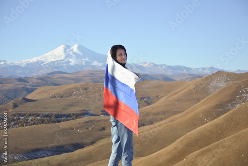Obraz na płótnie A young brunette woman stands against the backdrop of the snow-capped Mount Elbrus, looking at the camera, a Russian flag covers her shoulders. Tricolor against the backdrop of snow-capped Mount w salonie
