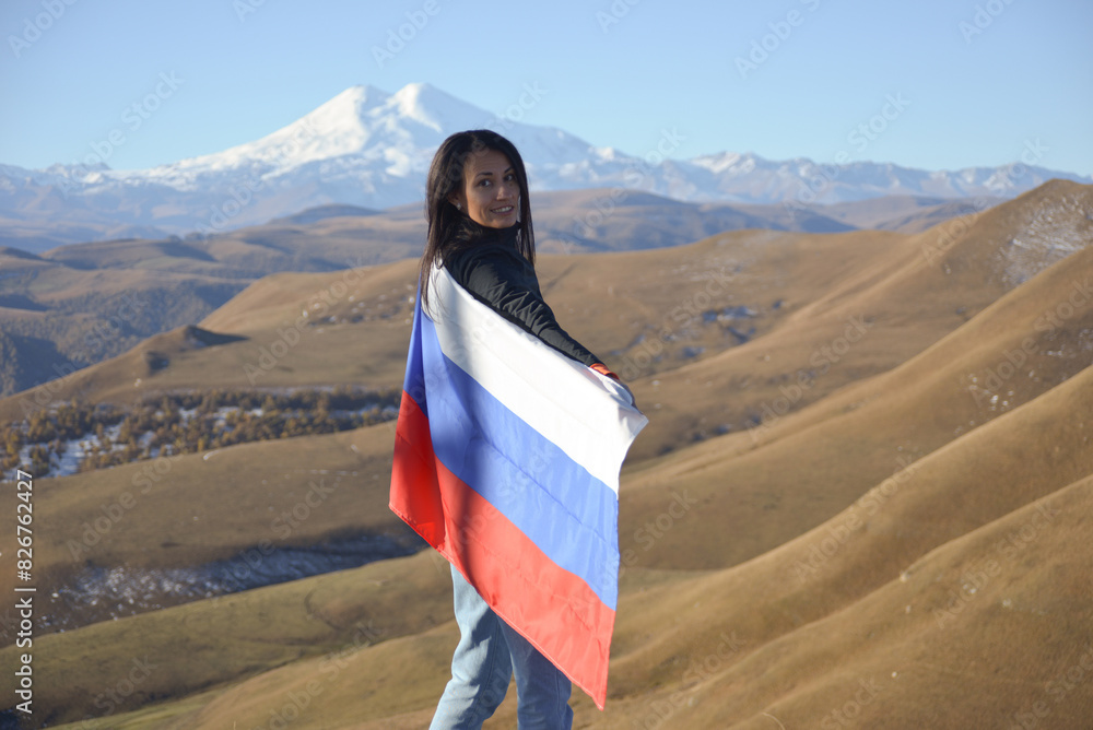 A young brunette woman stands against the backdrop of the snow-capped Mount Elbrus, looking at the camera, a Russian flag covers her shoulders. Tricolor against the backdrop of snow-capped Mount