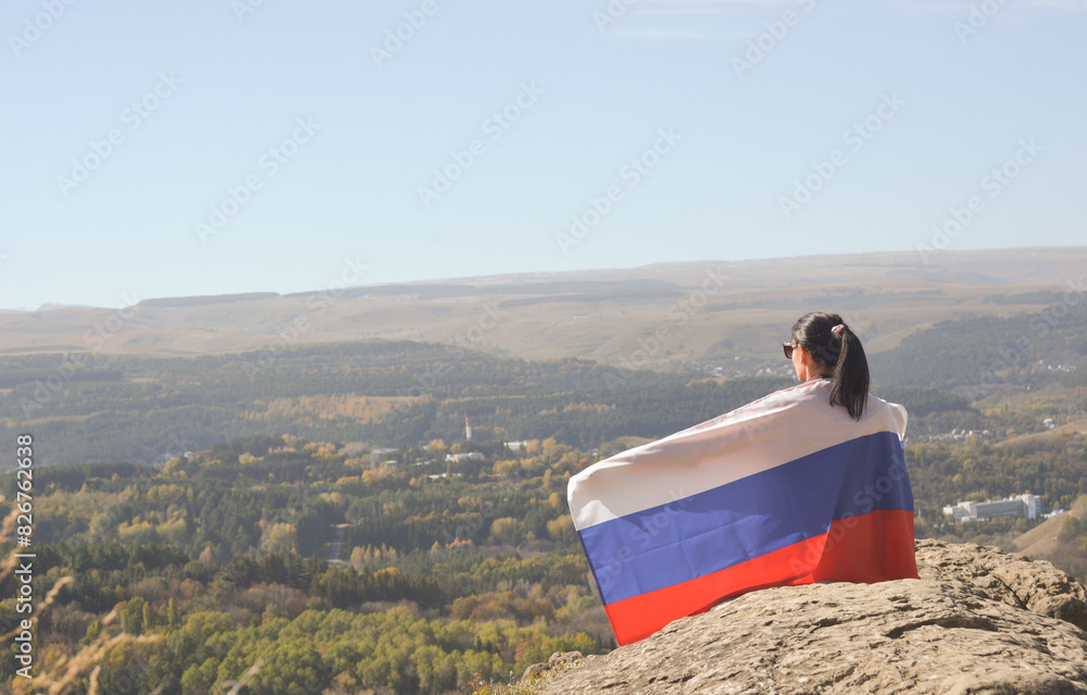 A young brunette woman sits on a stone and looks at the mountains around, her shoulders are covered with a Russian flag
