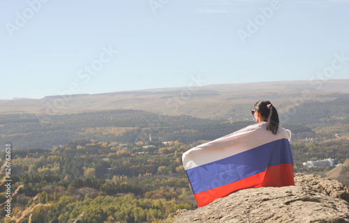 Obraz na płótnie A young brunette woman sits on a stone and looks at the mountains around, her shoulders are covered with a Russian flag w salonie